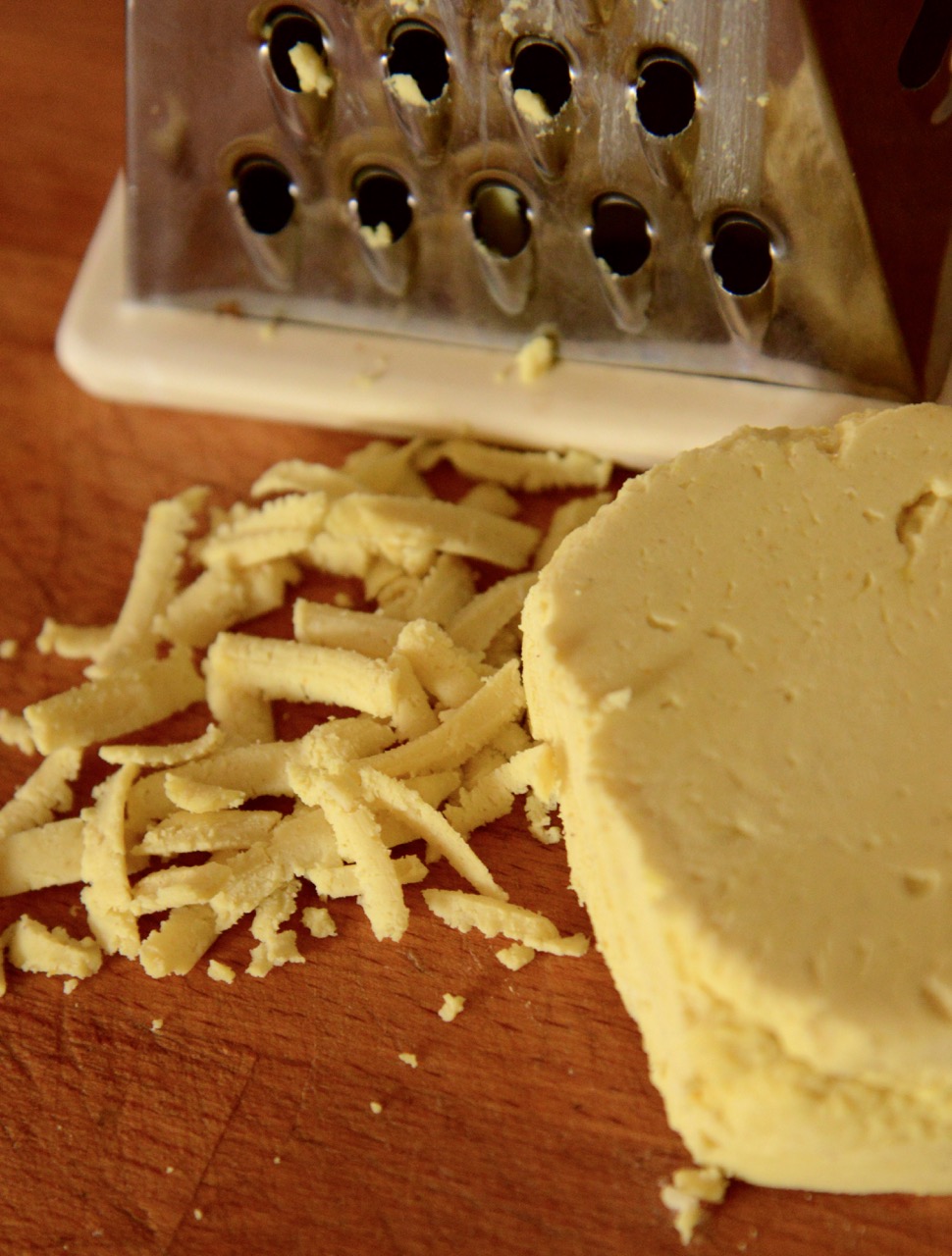 A block of plant-based cheddar cheese presented on a wooden cutting board, shown with shards of grated planted-based cheese, next to the metal grater used to grate it.