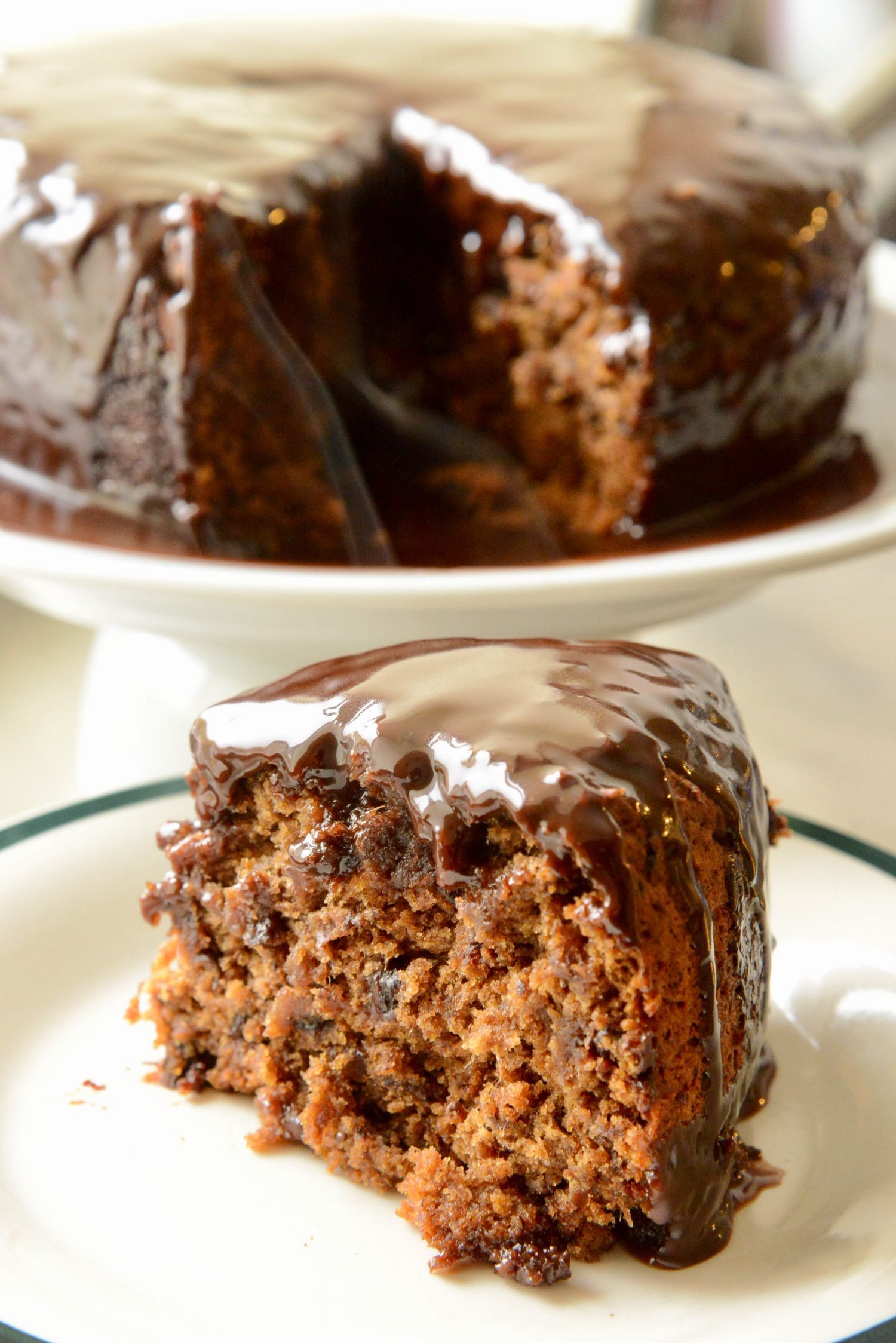A slice of sticky date pudding cake doused in whisky chocolate sauce presented on a plate in front of the cake on a pedestal.