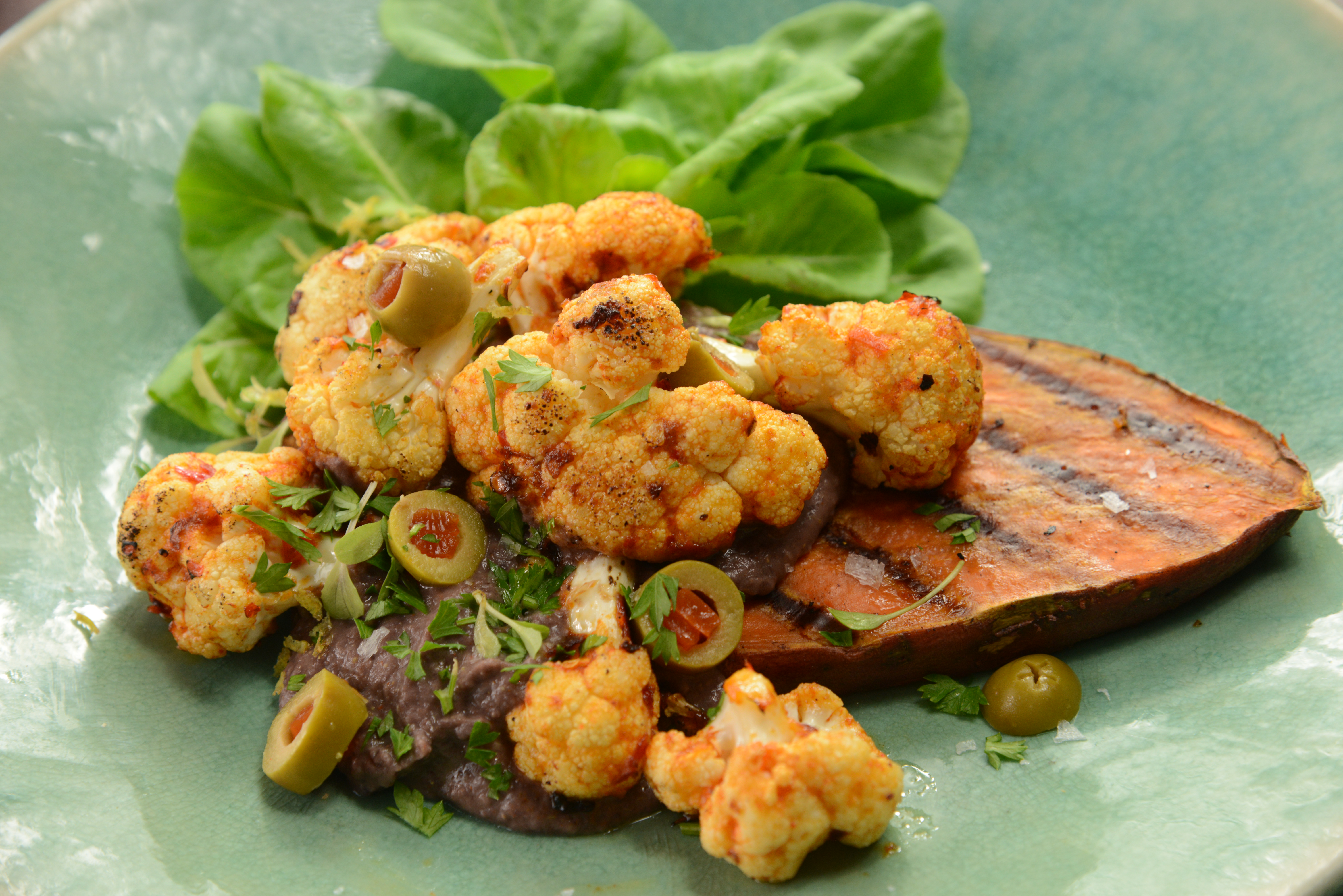 A plate of Kumara Steaks with Harissa Roasted Cauliflower and Black Turtle Bean Hummus served with Olives and Lettuce