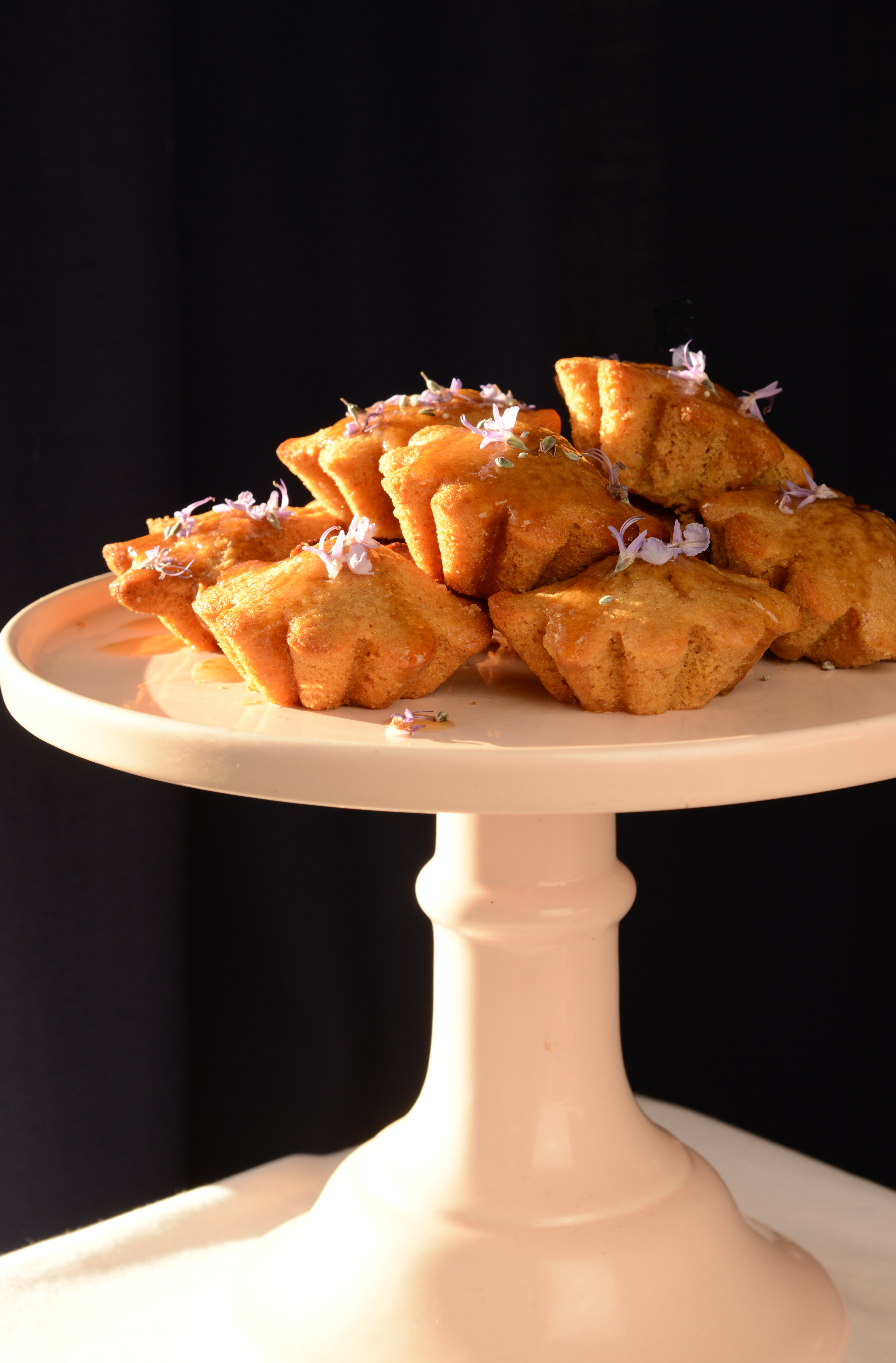 A cake stand displaying Kawakawa Tea Cakes glazed with syrup and garnished with rosemary flowers.