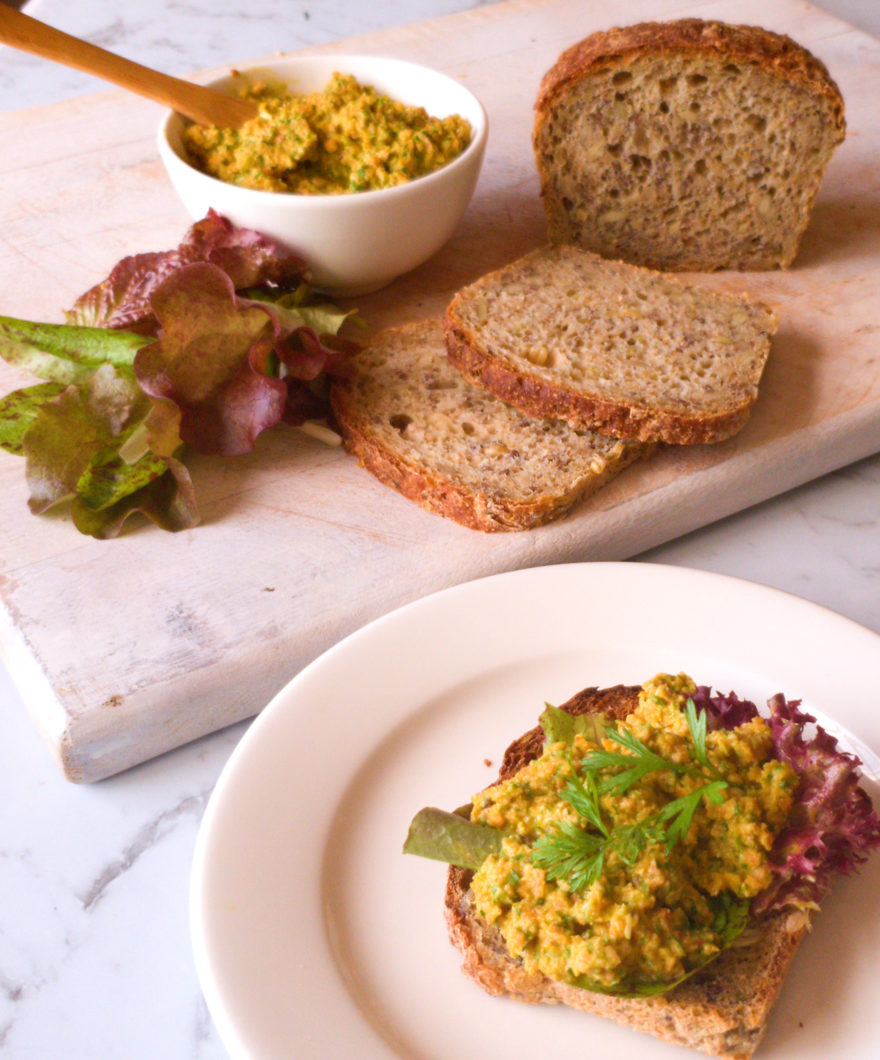 A bread board with slices of whole grain bread, lettuce leaves and a bowl of cashew and carrot top pesto beside an open sandwich