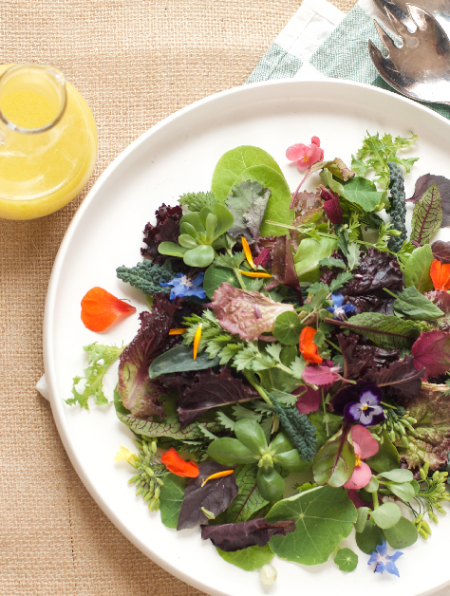 White platter of freshly picked seasonal salad greens with a jug of vinaigrette